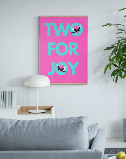 Living room with a pink poster reading 'TWO FOR JOY', a potted plant, and a gray sofa.