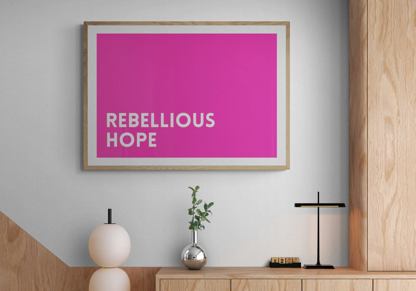 Modern interior with a bold bright pink framed poster with 'Rebellious Hope' wording on a light gray wall.