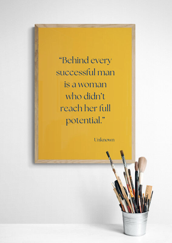 Framed quote "behind every successful man is a woman who didn't reach her full potential" on a yellow background with paintbrushes in a cup below.
