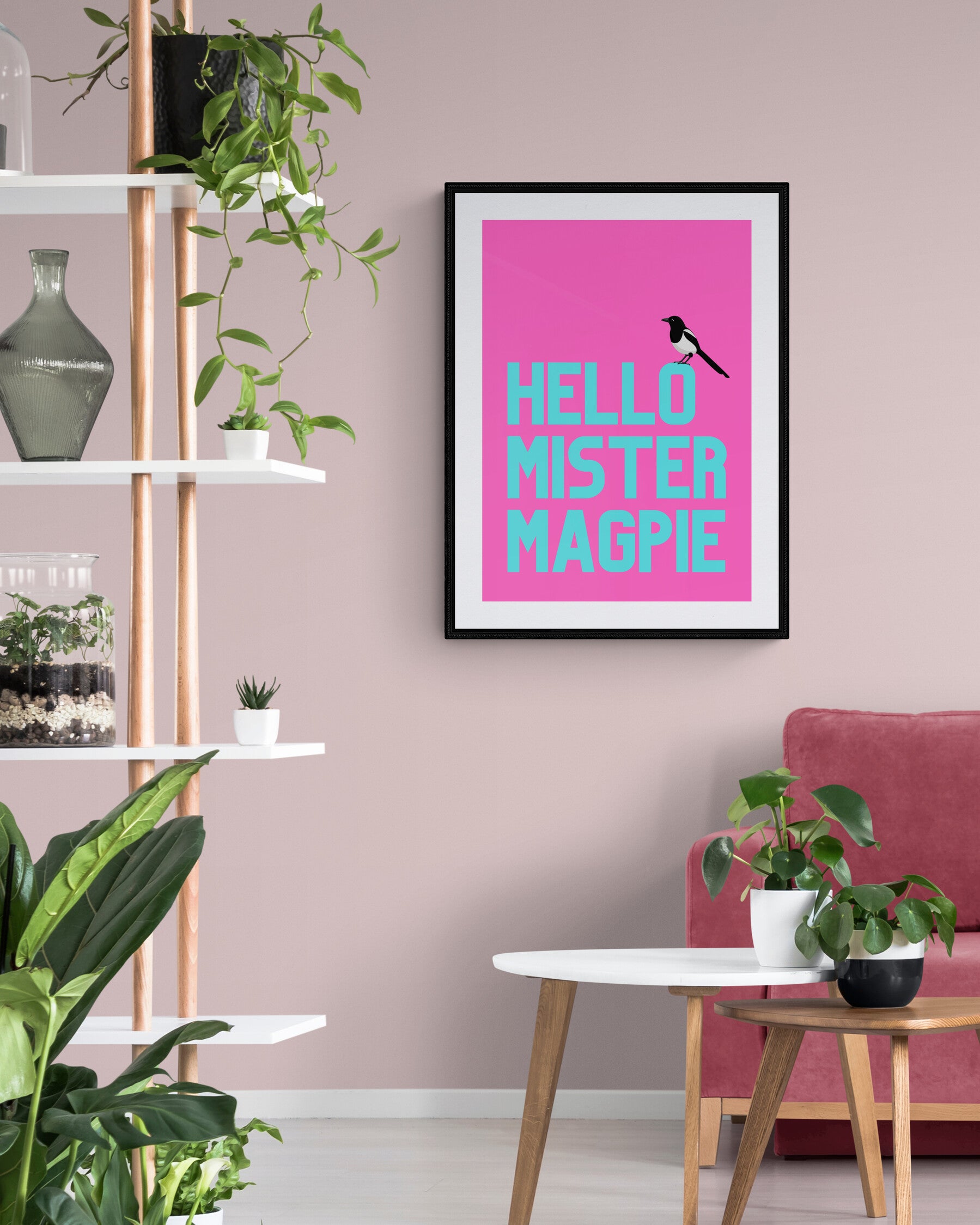 A vibrant wall art print with the phrase 'Hello Mister Magpie' featuring a black and white magpie on a bright pink background, displayed in a room with shelves and a pink armchair.