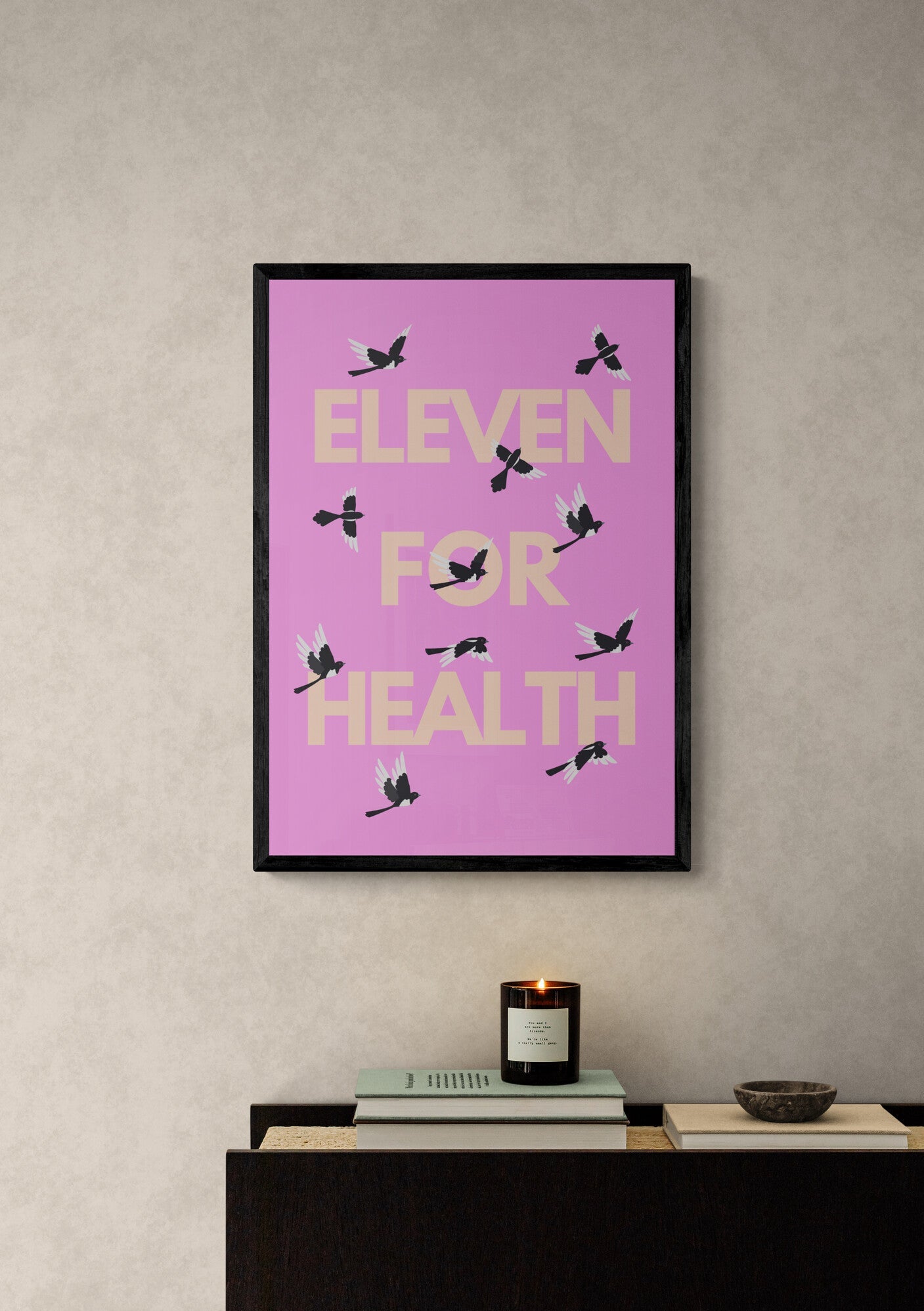 Framed poster with 'Eleven for Health' text on a pink background, displayed on a wall above a desk with eleven magpies positioned across the wording. 