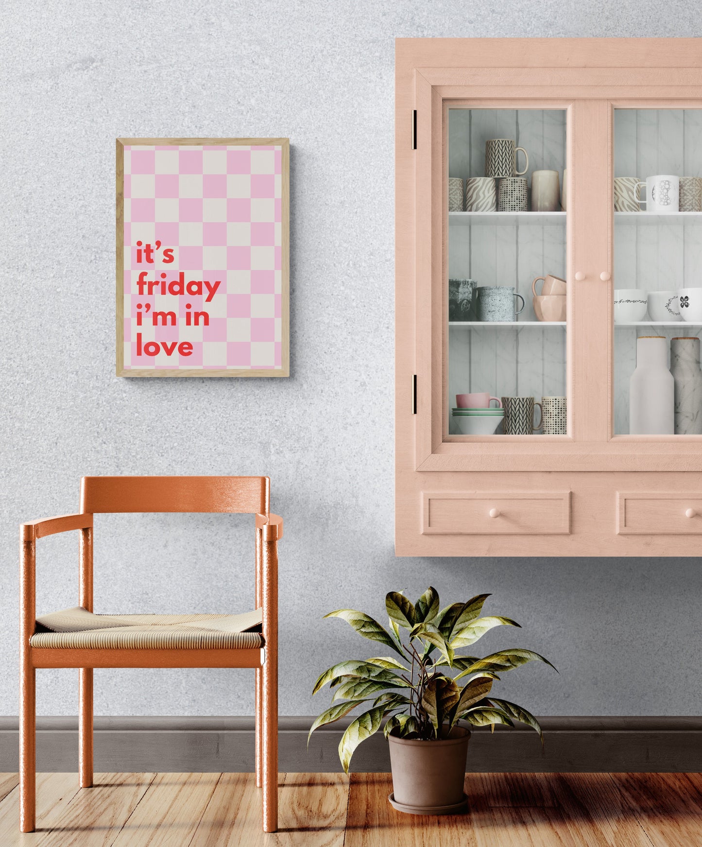 A framed print with a pink checkered background and the text 'it's friday i'm in love' displayed prominently, hanging on a wall above a chair and below a shelf with a plant.