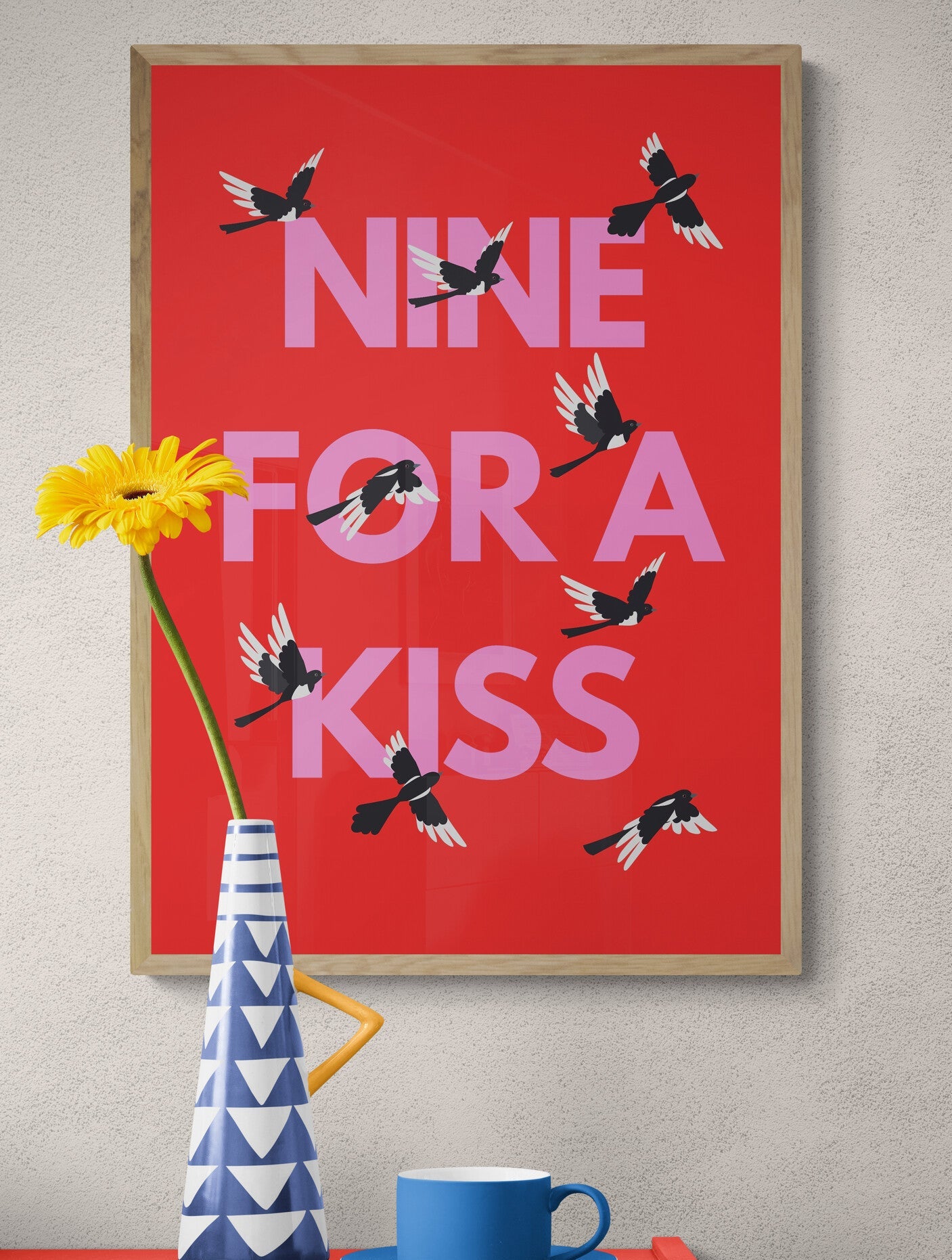 Framed poster with 'Nine for a Kiss' text and birds on a red background, placed above a red surface with a vase and cup.