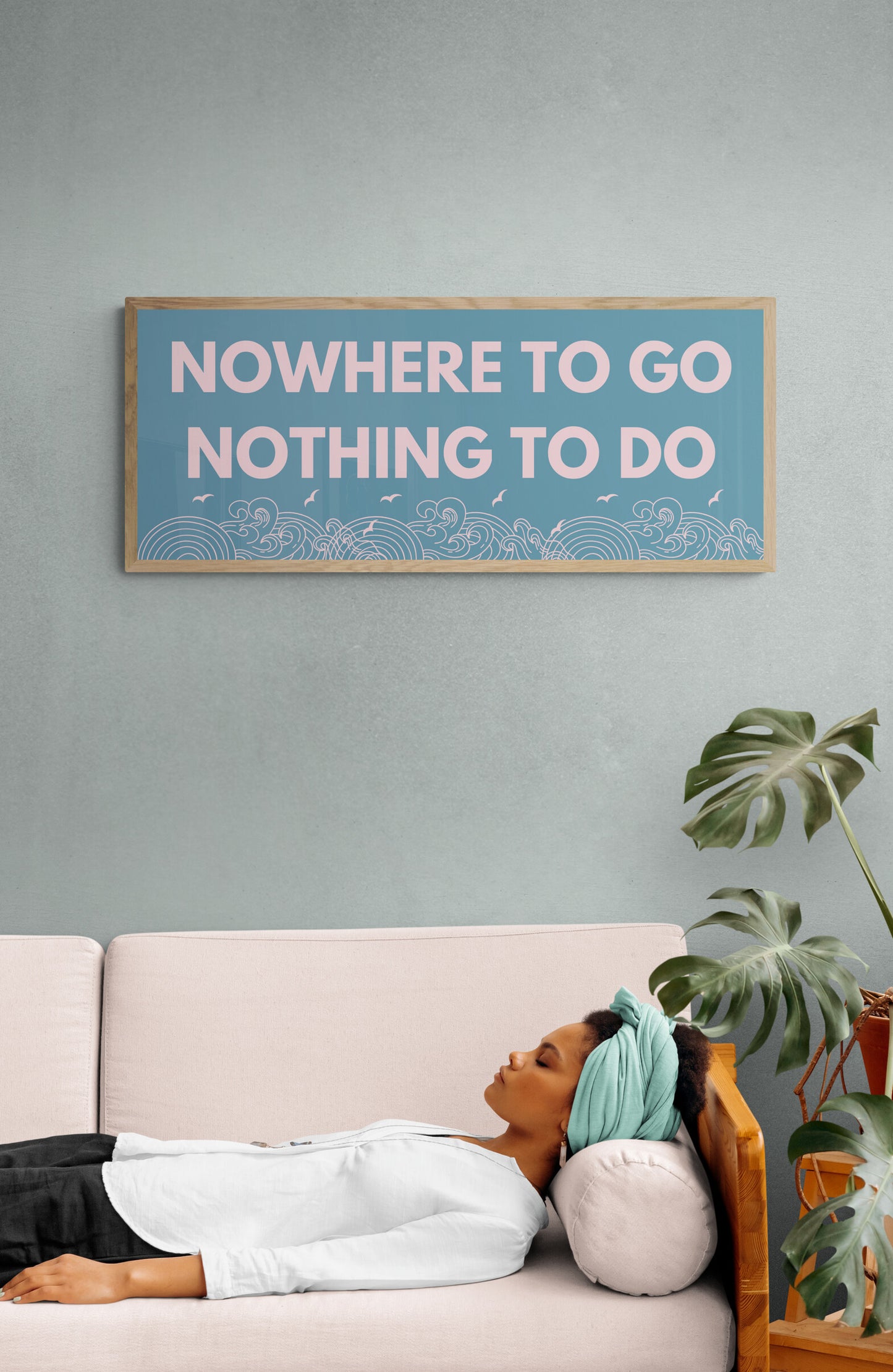 A wall art print with the text 'NOWHERE TO GO NOTHING TO DO' displayed in capital letters on a blue background, hanging on a wall above a sofa with a person lying on it.