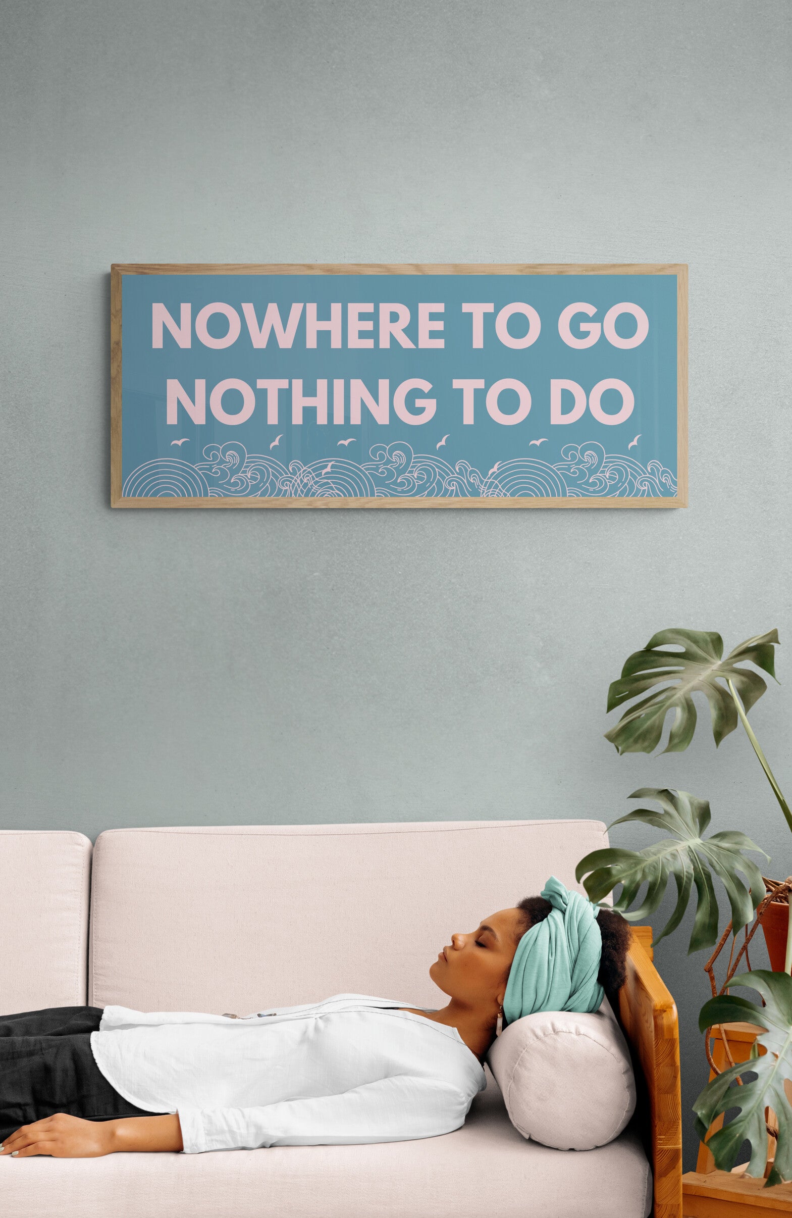 A wall art print with the text 'NOWHERE TO GO NOTHING TO DO' displayed in capital letters on a blue background, hanging on a wall above a sofa with a person lying on it.
