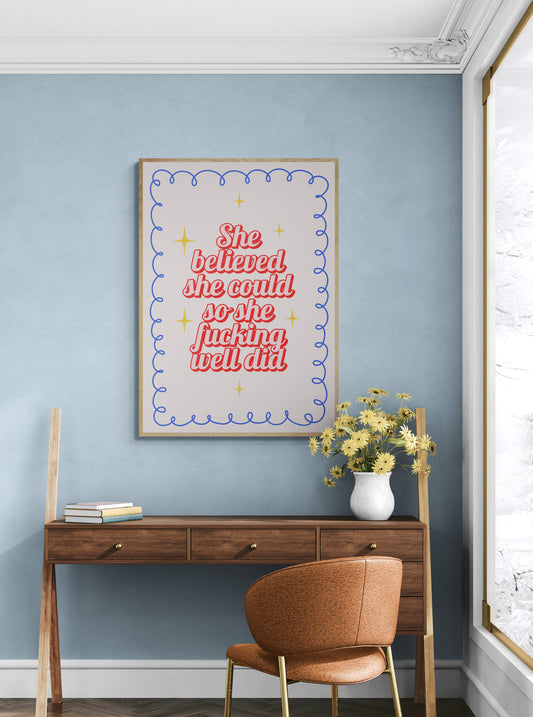 Wooden desk with a chair against a blue wall with a motivational poster saying "she believed she would so she fucking well did".